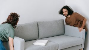 Two people carefully lifting a grey sofa in a plain, empty living room during a house move